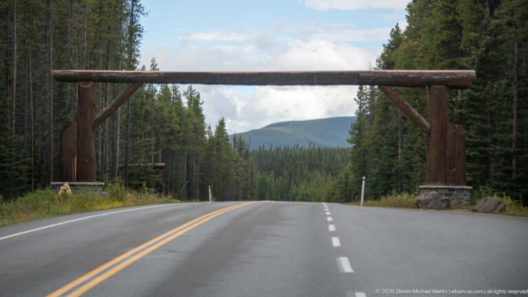 Large timber gateway spans an empty two-lane road descending through conifer forest toward low mountains.