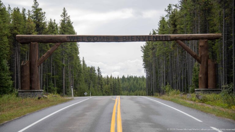Timber arch marked “WELCOME • BIENVENUE” spans a wet two-lane road through dense conifer forest.