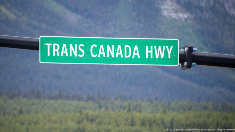 Green sign reading “TRANS CANADA HWY” mounted against a blurred forested mountain background.