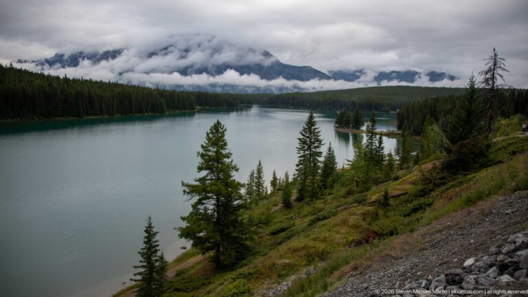 Long blue-green lake bordered by conifer forest and steep banks beneath cloud-covered mountains.