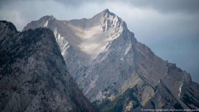 Steep pale limestone peak with sharp summit and gullied face rises beneath dark storm clouds.