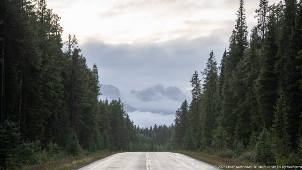 Wet two-lane forest road leads straight toward mist-covered mountain peaks under a gray sky.