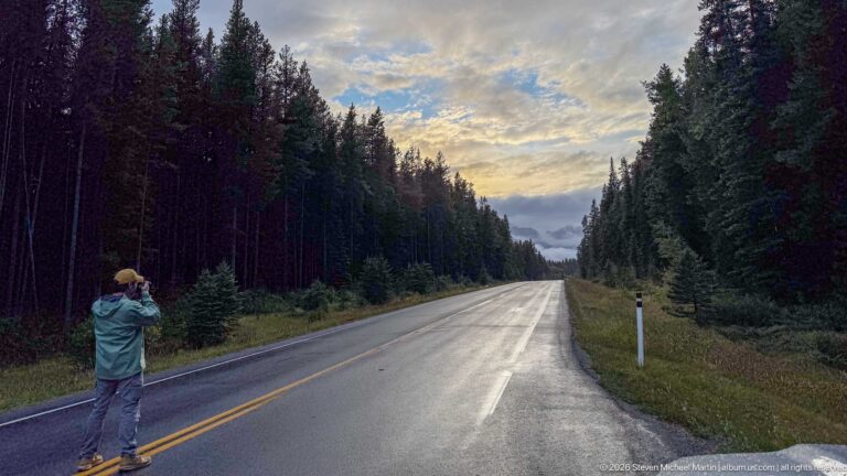 Photographer stands on the centerline of a wet forest road beneath a brightening cloud-filled sky.