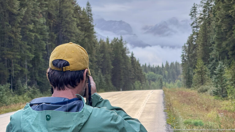 Person in a cap and rain jacket photographs a straight forest road leading toward cloudy mountains.