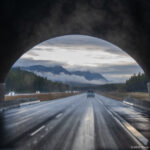 Dark wildlife-crossing arch frames wet divided highway lanes and distant forested mountains.