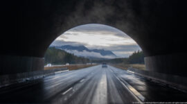 Dark wildlife-crossing arch frames wet divided highway lanes and distant forested mountains.