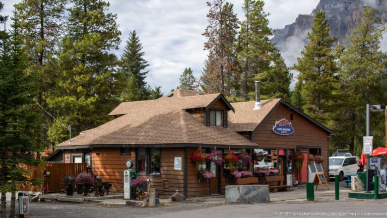 Wood-sided lodge office with flowers, signs, vehicles, and a limestone mountain wall behind.