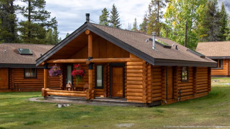 Brown log cabin with gabled porch, hanging flowers, and neighboring lodge buildings among trees.