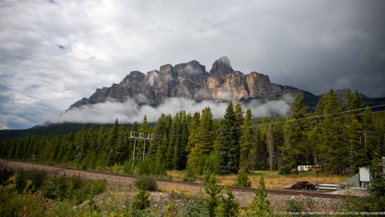 Jagged limestone ridge rises above low cloud, conifer forest, railway tracks, and utility lines.