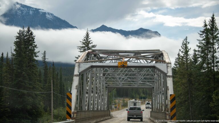 Vehicles pass through a gray steel truss bridge surrounded by conifers and low mountain clouds.