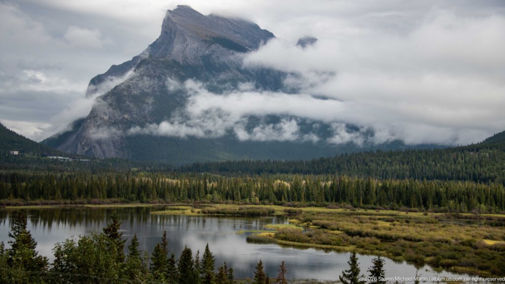 Cloud-wrapped mountain rises beyond conifer forest and reflective marsh water in the Bow Valley.