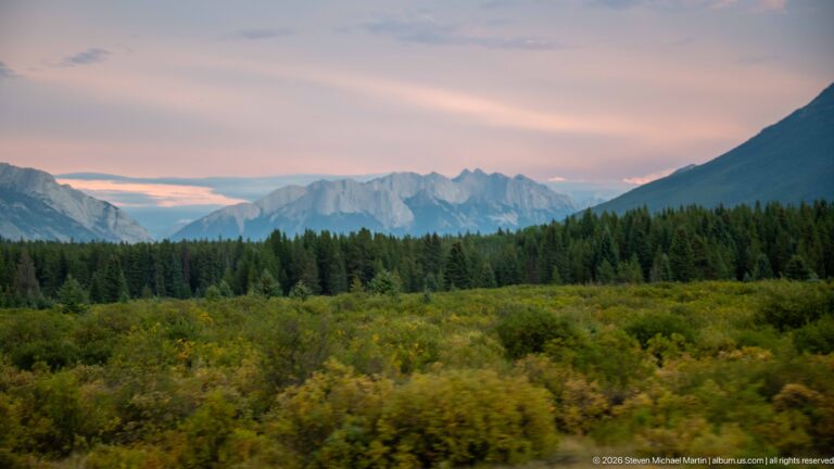 Layered shrubs and conifer forest lead toward distant blue-gray mountain peaks beneath a pink evening sky.