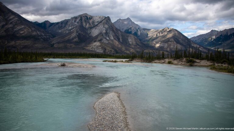 Turquoise river curves around pale gravel bars below rugged gray mountains and scattered conifers.
