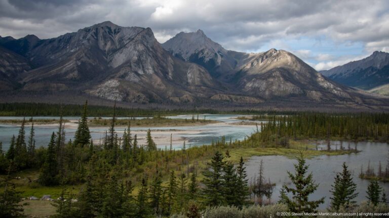 Multiple turquoise river channels and conifer islands cross a broad floodplain beneath rugged mountains.