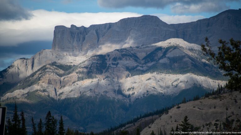 Massive layered limestone mountain illuminated by broken sunlight beneath dark storm clouds.