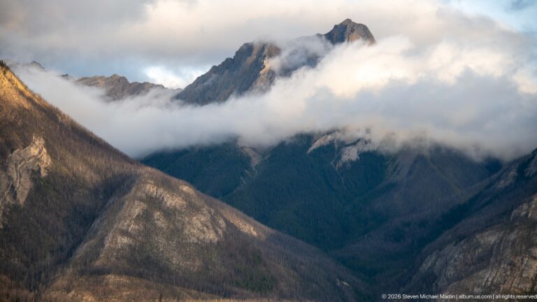 Jagged mountain summit lit above a band of low cloud and dark forested slopes.