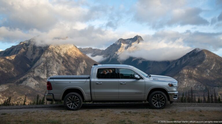 Silver pickup truck with beagles inside, parked before cloud-wrapped Rocky Mountain peaks.