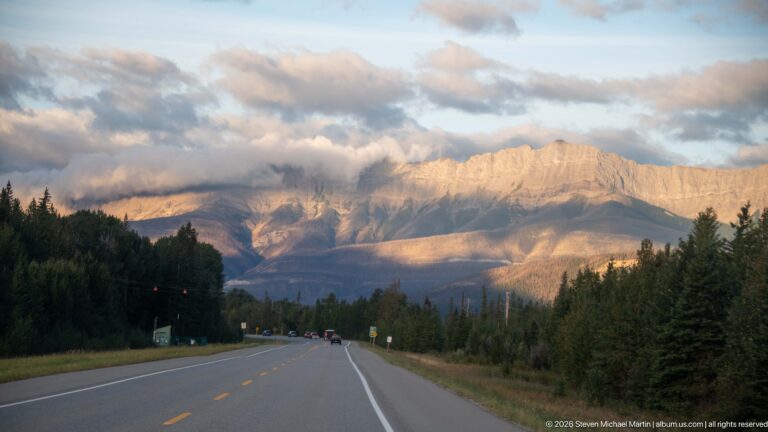 Two-lane highway leading toward a broad sunlit Rocky Mountain wall.