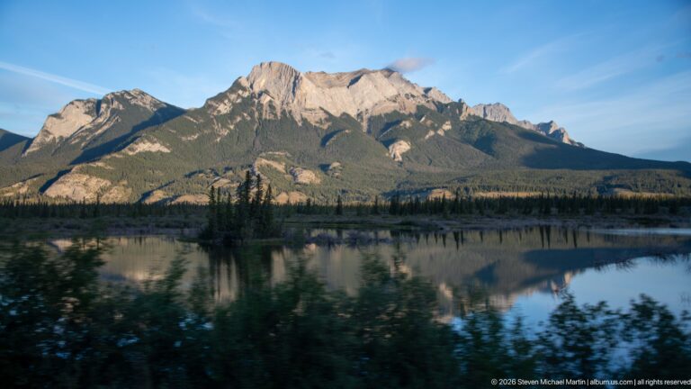 Rugged mountain massif and conifer forest reflected in calm blue water.