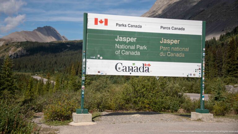Green bilingual Parks Canada sign for Jasper National Park with forested mountains behind.
