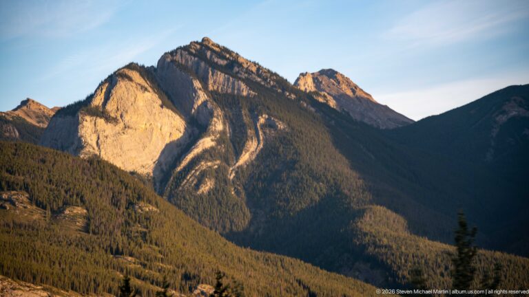 Sunlit pale mountain face rising above dark conifer forest and shadowed slopes.