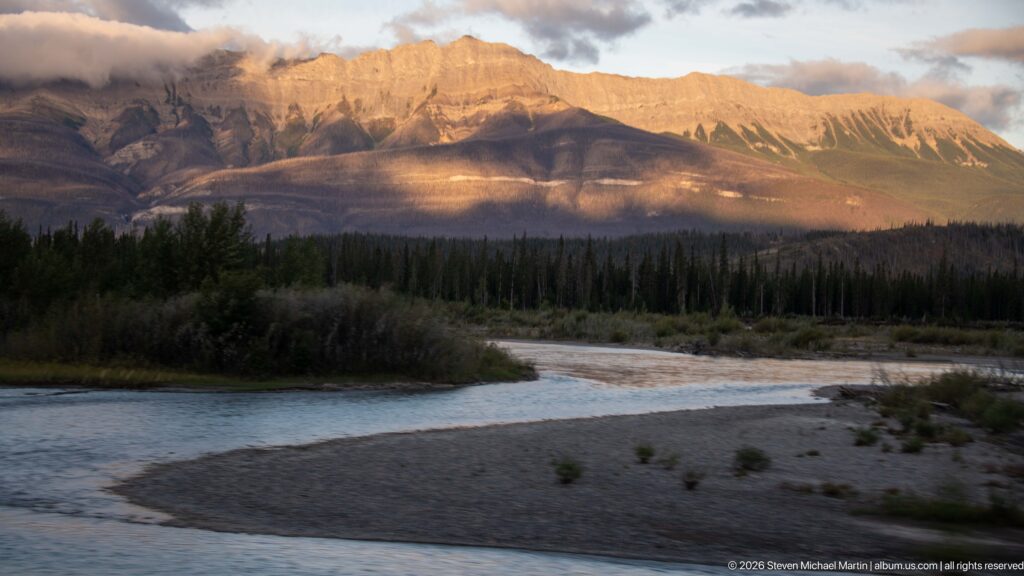 Shallow river channel curving around a gravel bar below a sunlit mountain wall.