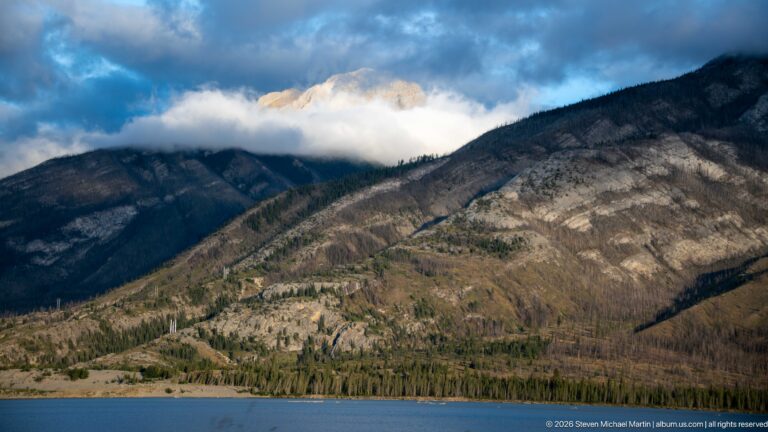 Blue water below steep rocky and forested mountain slopes with clouds around the summit.