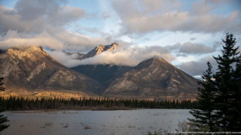 Broad gravel flats below forested mountains and cloud-wrapped peaks.