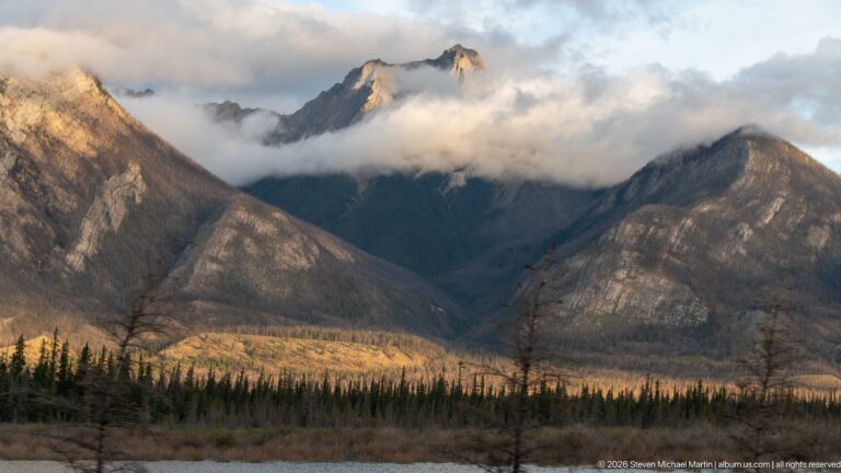 Cloud-wrapped mountain peaks above a dark conifer forest in warm evening light.
