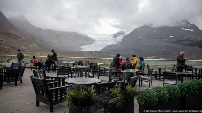 Visitors on an outdoor terrace with patio tables overlooking Athabasca Glacier and cloud-covered mountains.