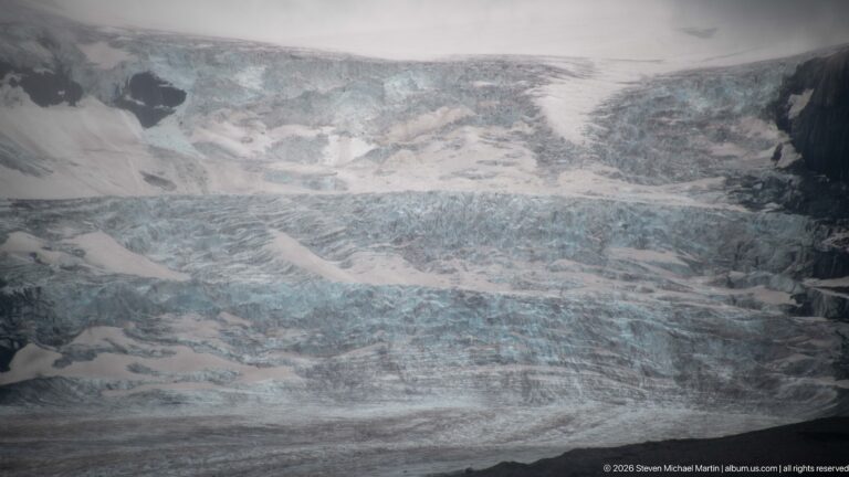 Telephoto view of Athabasca Glacier with fractured blue-white ice, deep crevasses, seracs, snow, and debris bands.