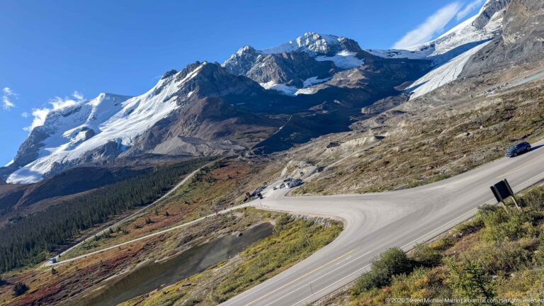 Winding Icefields Parkway crossing an open mountainside beneath snowfields, glacier ice, and steep rocky peaks.