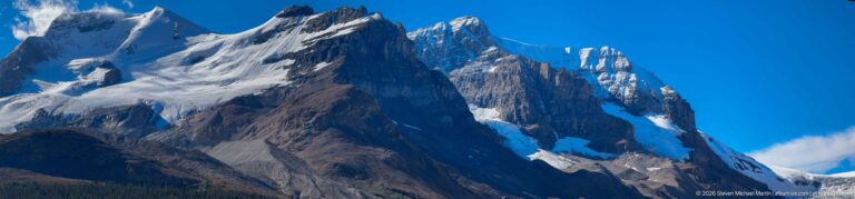 Jagged dark mountain summit with steep cliffs, snowfields, and small ice patches beneath a vivid blue sky.