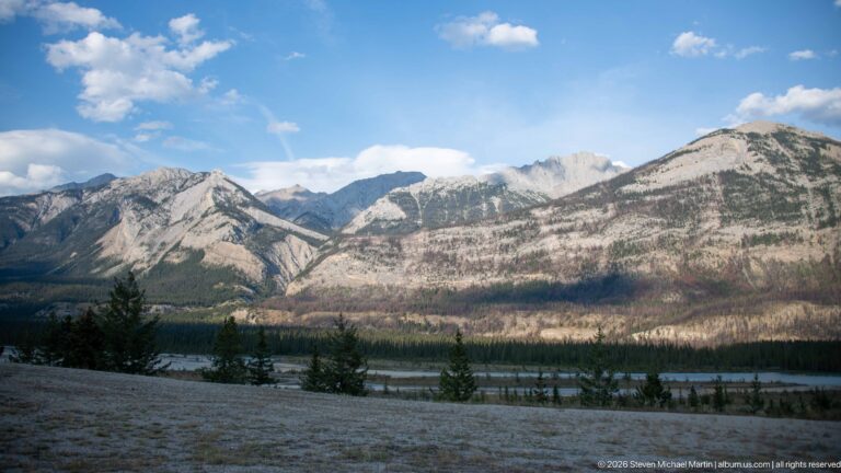 Pale layered mountains rise beyond a valley with sparse fire-affected forest and a braided river corridor.