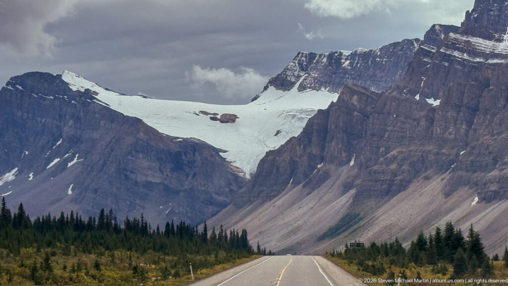 Two-lane Icefields Parkway leading toward steep layered cliffs and a broad high snow-and-ice field beneath clouds.