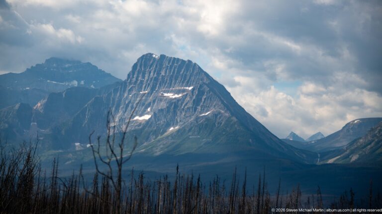 A steep dark mountain with strongly tilted rock layers rises beneath broken clouds above bare burned tree trunks.