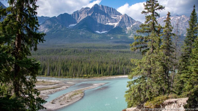A turquoise river curves around gravel bars below conifer forest and steep mountain peaks.