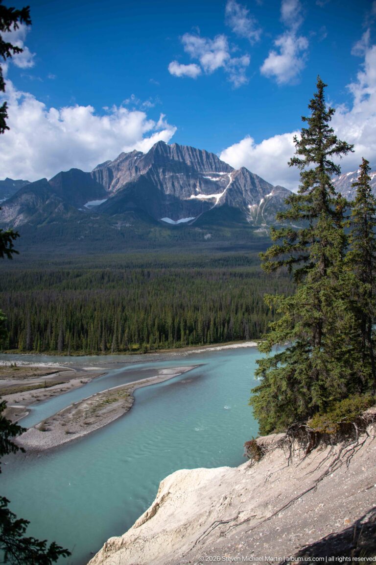 Blue-green water flows past pale sediment bars with forest and a long layered mountain ridge beyond.