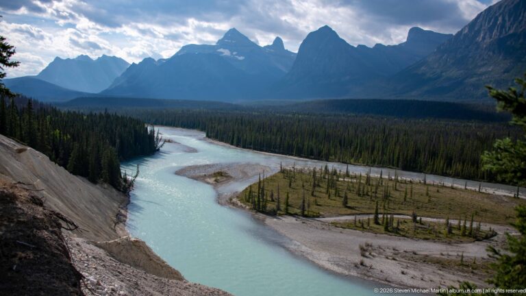 A pale turquoise river bends around gravel islands and sparse trees beneath jagged mountain silhouettes.