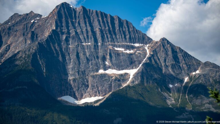 A massive dark mountain face displays steep parallel rock bands, gullies, and isolated snow patches.