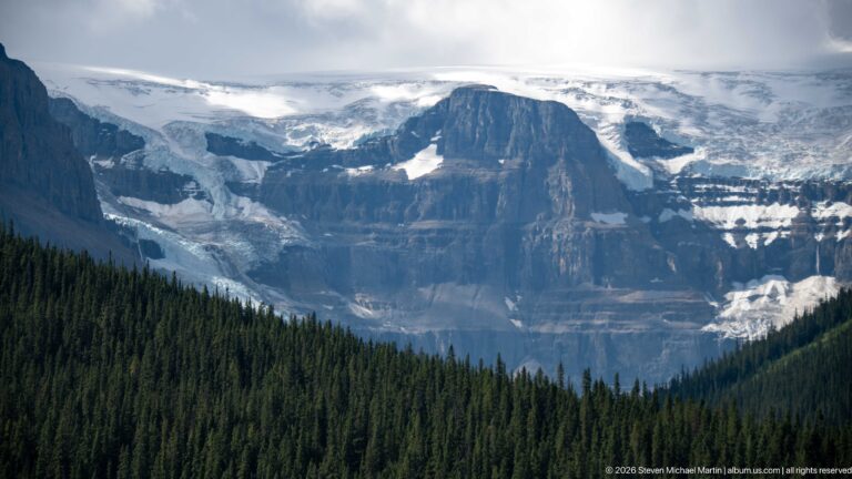 A broad glacier and snowfield descend around a dark rock buttress above dense conifer forest.