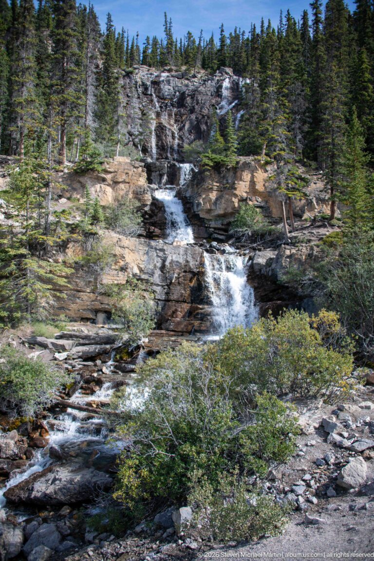 A stream spreads over multiple rock ledges amid conifers, shrubs, and exposed stone.