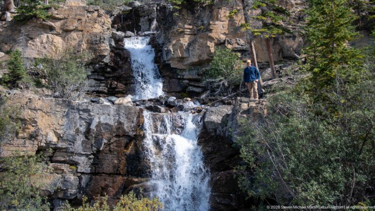 A visitor stands on a ledge beside a two-tier waterfall dropping through layered rock.