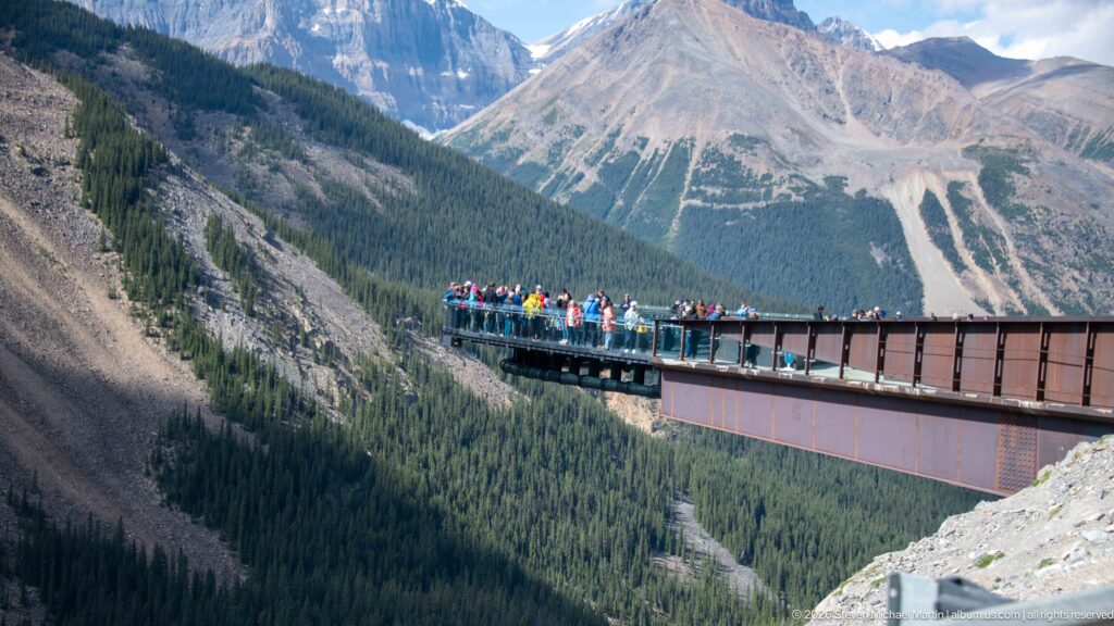A crowd stands on a projecting glass-railed observation platform above a deep forested mountain valley.