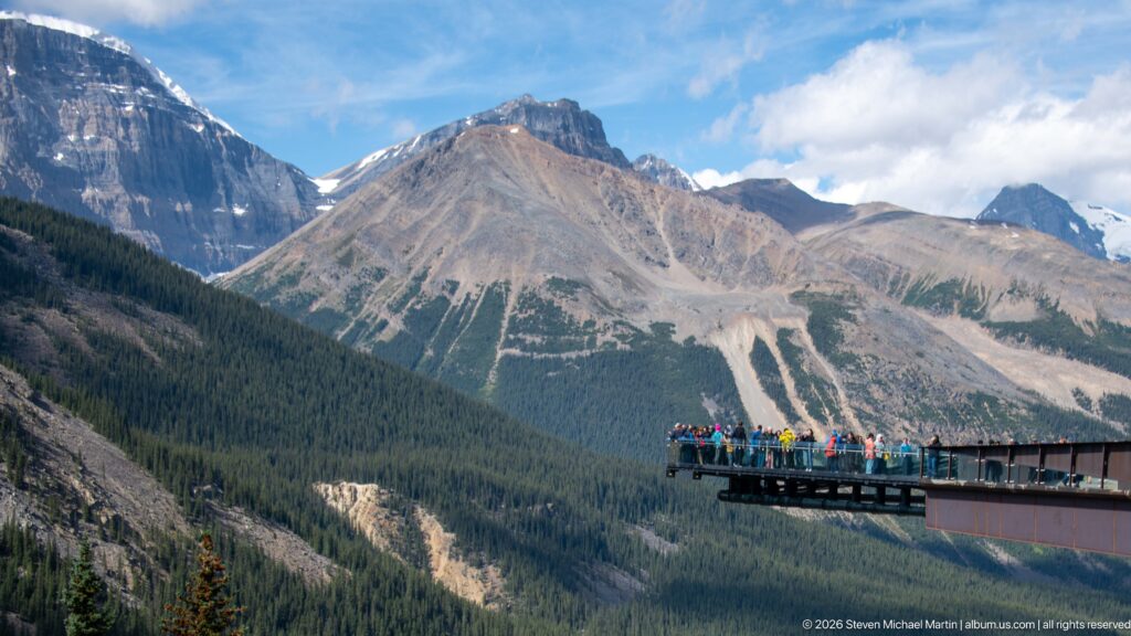 A glass-railed platform crowded with visitors projects from the right edge toward a broad mountain valley.