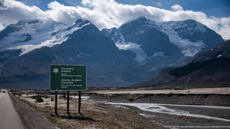 A green Columbia Icefield sign stands beside the road with braided meltwater channels and glacier-covered mountains beyond.