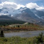 Glacier-covered peaks rise above a small pond, low shrubs, conifers, and a winding mountain road.