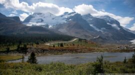 Glacier-covered peaks rise above a small pond, low shrubs, conifers, and a winding mountain road.
