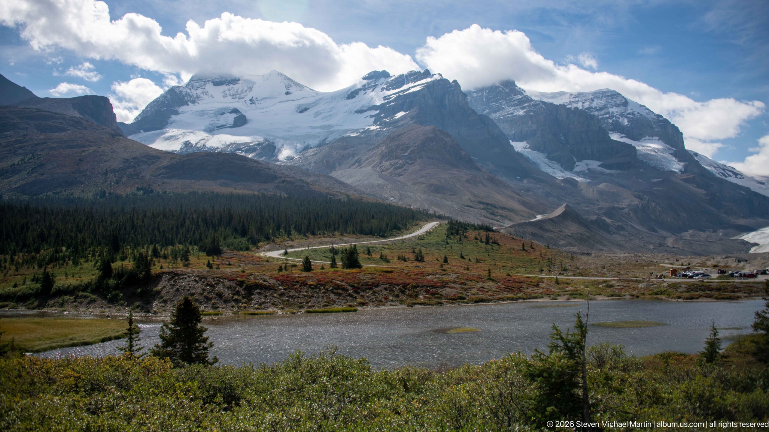 Glacier-covered peaks rise above a small pond, low shrubs, conifers, and a winding mountain road.