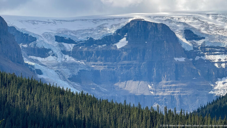 Glacier ice and snowfields surround a massive layered rock buttress above dense conifer forest.